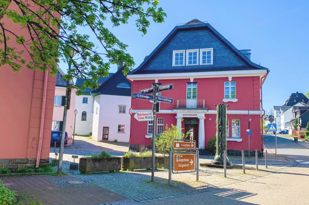 Auf dem Bild ist ein dunkelrotes Haus. Es ist die Kornbrennerei Krugmann. Vor dem Haus stehen Schilder und ein Brunnen. Es ist ein sonniger Tag. Der Himmel ist blau