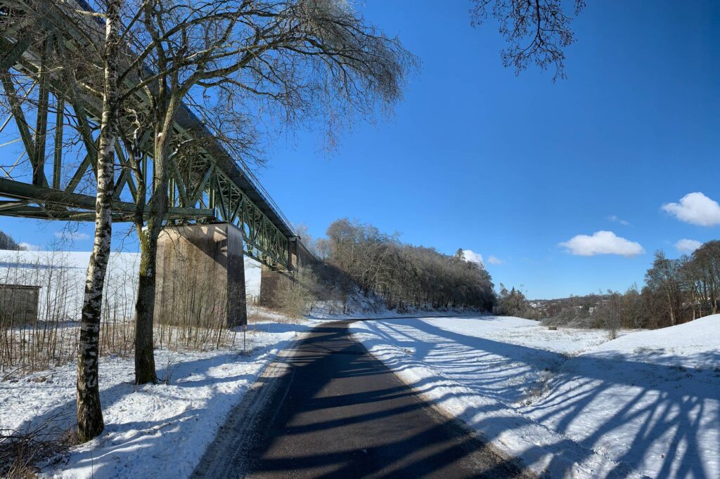 Auf dem Bild ist eine Straße zwischen Schneewiesen. Links steht eine große Brücke. Es ist die Fischbauchbrücke in Meinerzhagen.