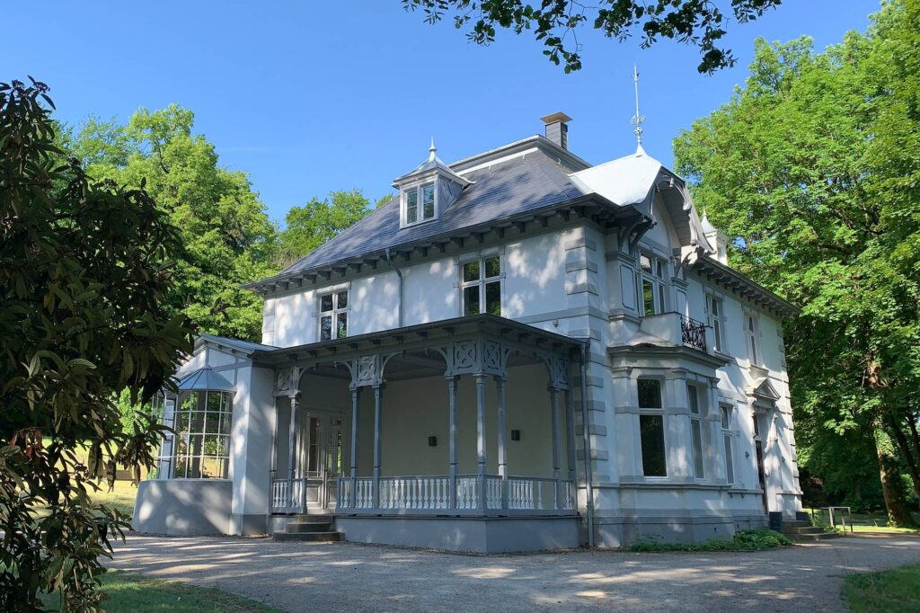 Auf dem Bild ist eine weiß-graue Villa. Sie steht zwischen grünen Bäumen. Es ist die Villa im Volkspark Meinerzhagen. Sie heißt Villa Schmiemicke
