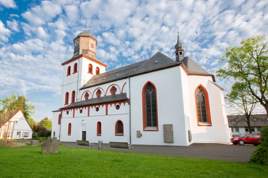 Auf dem Bild ist eine große Kirche. Sie ist weiß mit dunkelroten Fenstern und zwei Türmen. Es ist die Jesus-Christus-Kirche in Meinerzhagen.