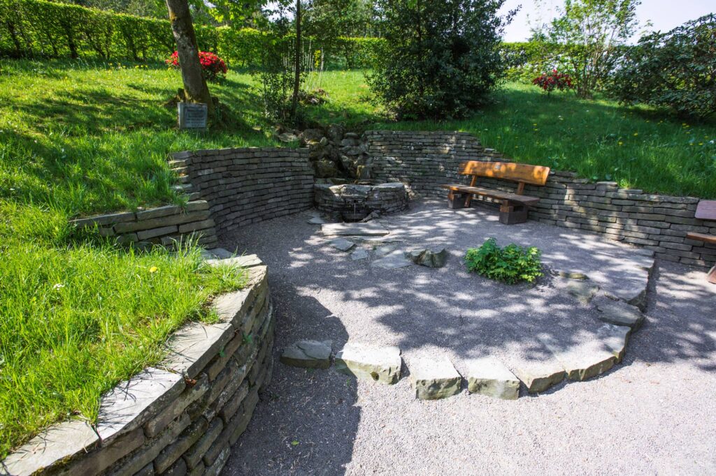 Auf dem Bild ist ein Platz mit Steinen. Es gibt eine Mauer und einen Brunnen. Der Brunnen ist die Volmequelle. Aus der Quelle fließen Wassertropfen. Es gibt auch eine Bank. Die Sonne scheint.